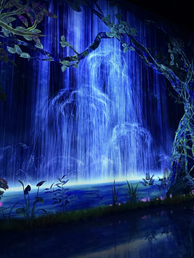 A vibrant, blue-lit waterfall cascades over rocks surrounded by lush greenery and foliage, with reflections shimmering on the water below, creating a magical, otherworldly scene.