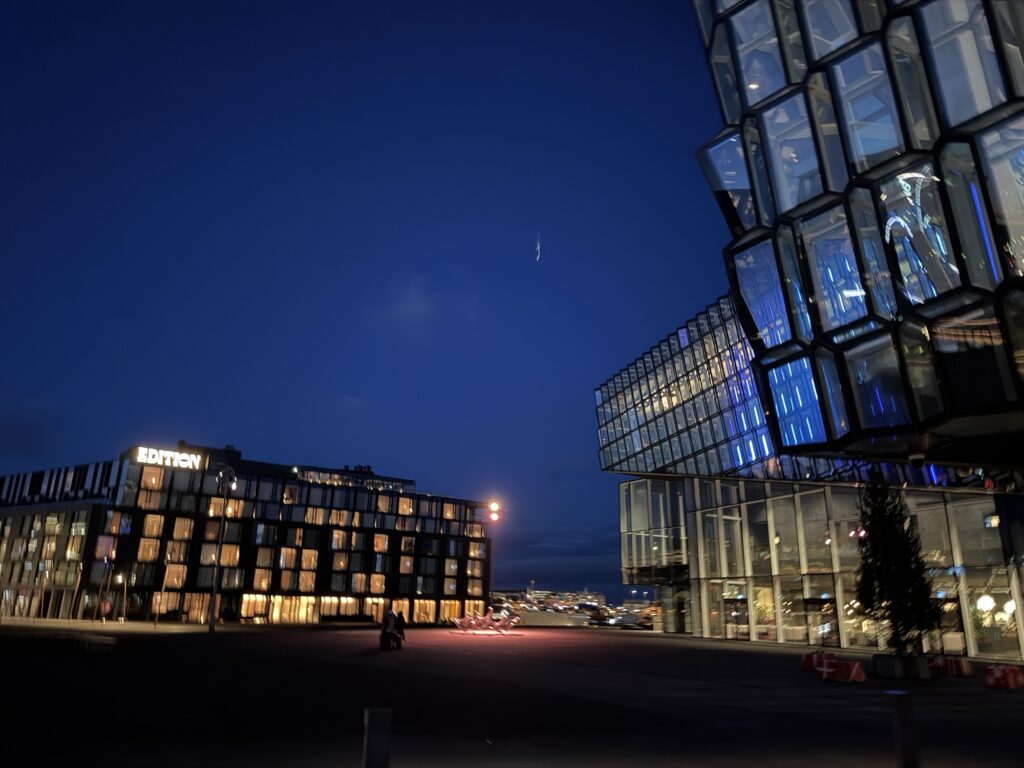 Modern glass buildings illuminated at night, with a clear dark blue sky above. A crescent moon is visible in the background, and a few people walk through the open plaza between the buildings.