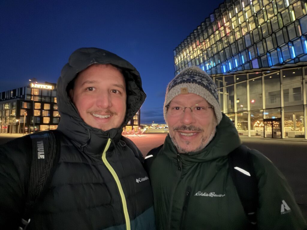 Two people in winter jackets and hats smile for a selfie outside at night in front of a modern glass building with colorful lights inside. Another illuminated building is visible in the background.