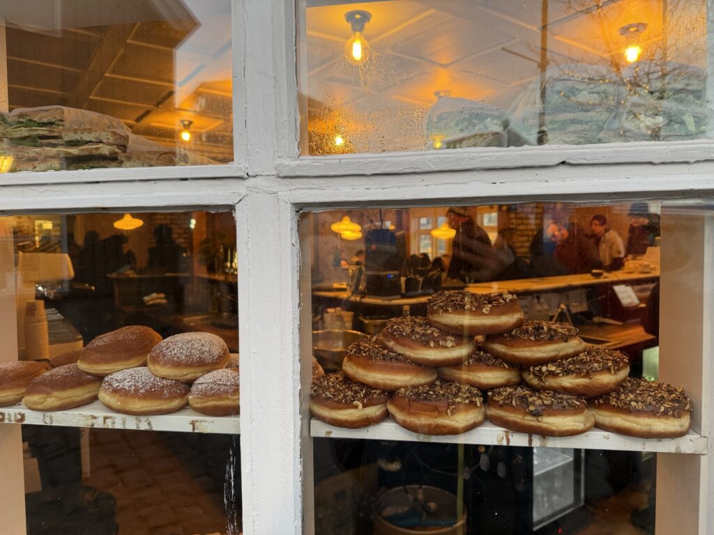 A bakery window displays shelves of powdered and glazed donuts. Inside, people wait at the counter under warm yellow lights, and reflections show the street outside.