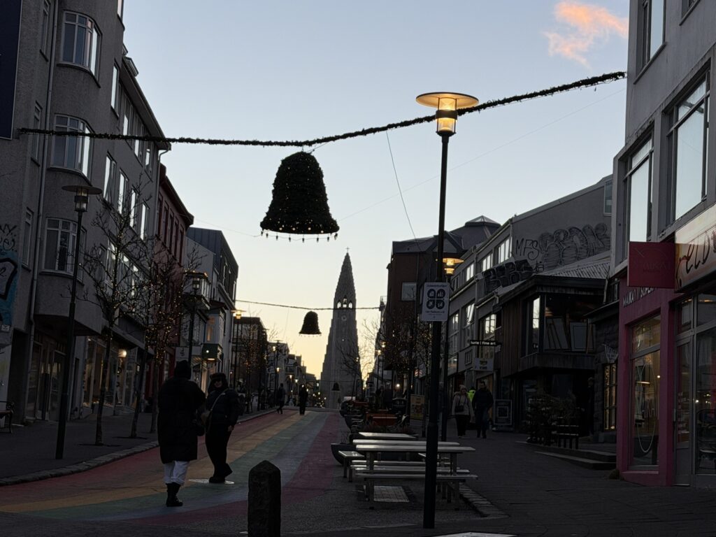 A street scene at dusk shows people walking under holiday decorations, with a large bell ornament hanging above. Buildings line both sides and a tall, pointed church rises in the background. Strings of lights are visible along the street.