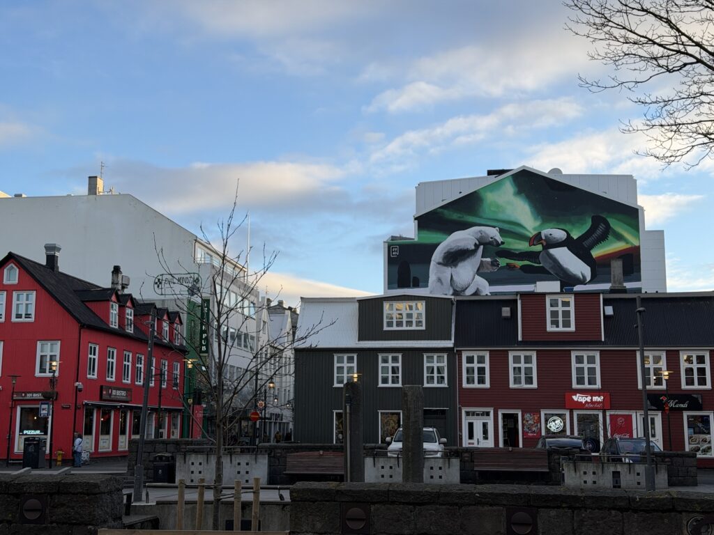 Colorful buildings with red and green facades stand beneath a large mural of two polar bears shaking hands under the northern lights on a white building. The sky is blue with scattered clouds, and a leafless tree is visible.