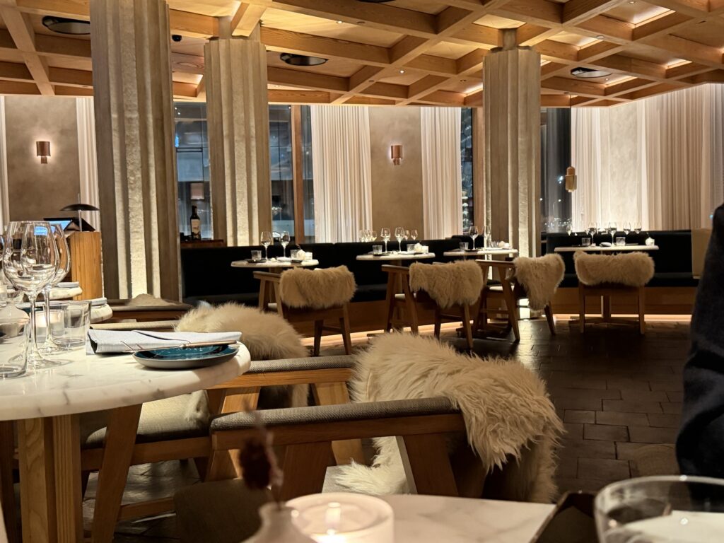 A warmly lit, modern restaurant interior featuring wooden tables and chairs draped with white faux fur throws, elegant table settings, tall curtains, and large windows showcasing an evening city view.