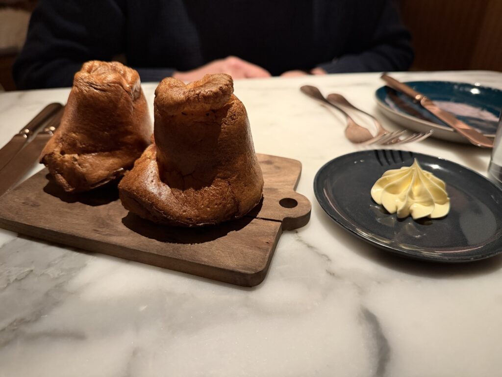 Two Yorkshire puddings on a wooden board beside a small plate with a swirl of butter, set on a marble table with cutlery and a blue plate in the background.
