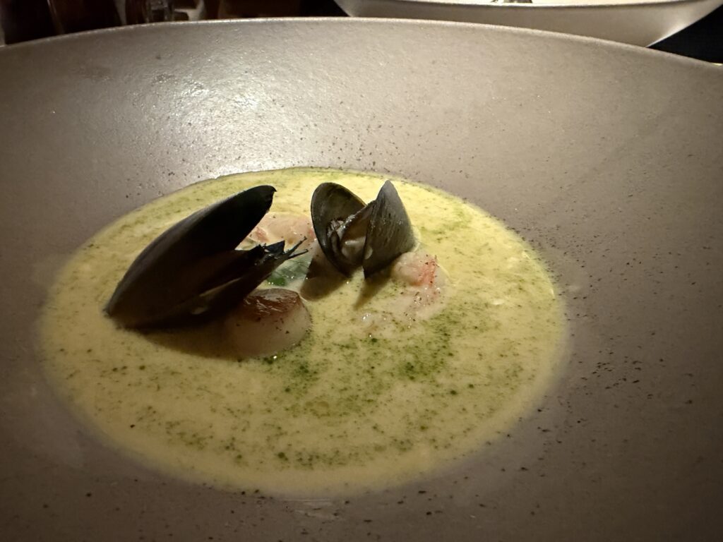 A shallow bowl of creamy yellow-green soup garnished with herbs, with clams in their shells and pieces of seafood visible in the center.