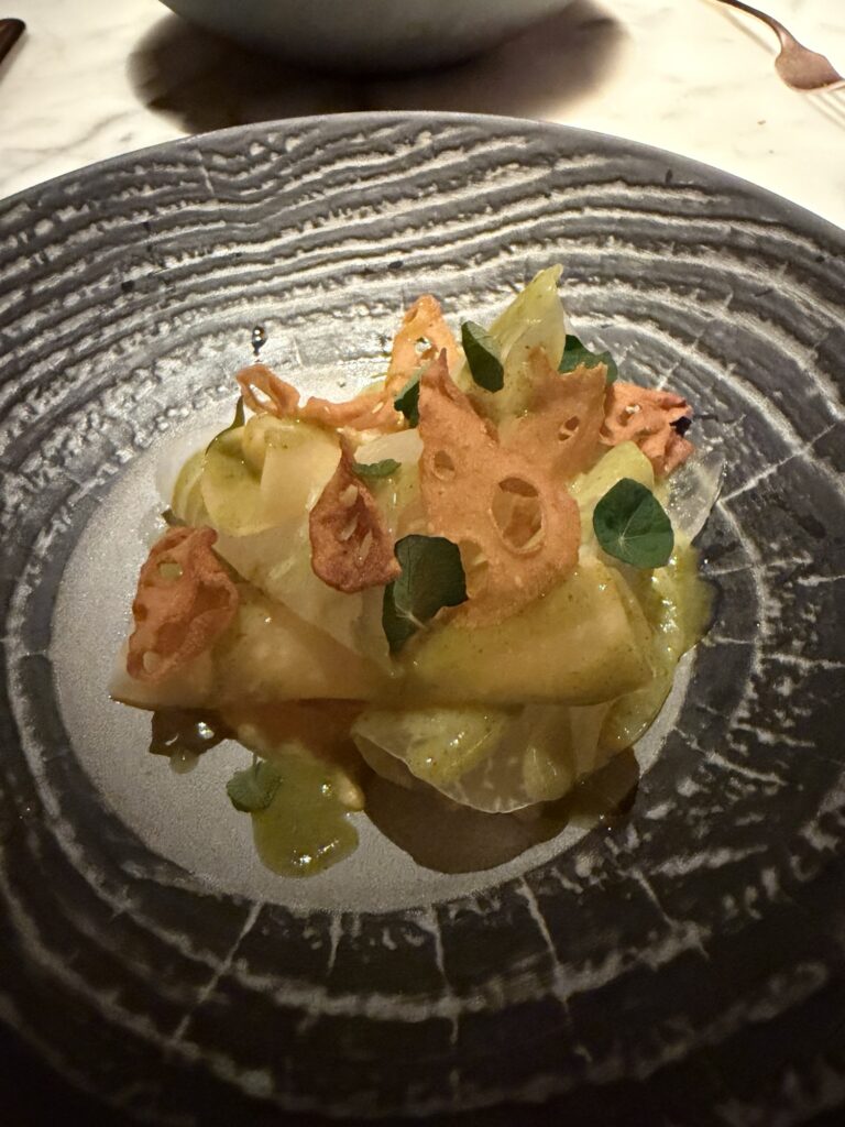 A dark ceramic plate with a gourmet dish featuring thinly sliced, translucent vegetables, golden crispy lotus root chips, green sauce, and small fresh greens, all artfully arranged on a textured surface.