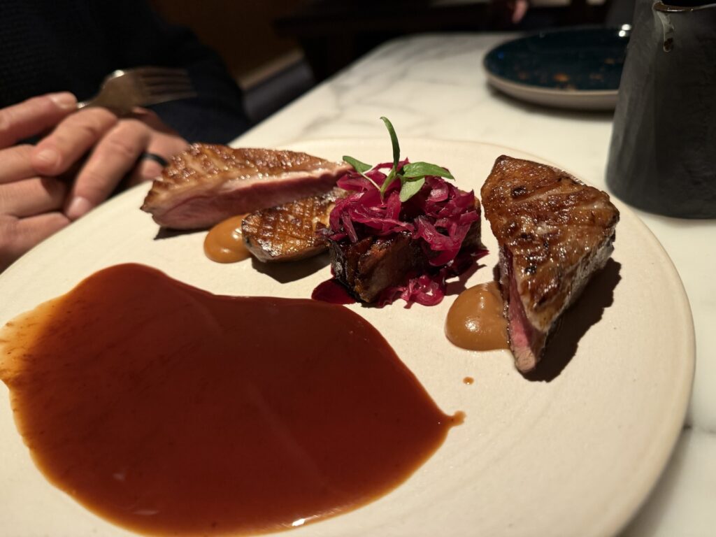 A plate with three slices of seared lamb, red cabbage slaw, dollops of brown sauce, and a pool of reddish-brown sauce. A person’s hands with a fork are visible in the background.