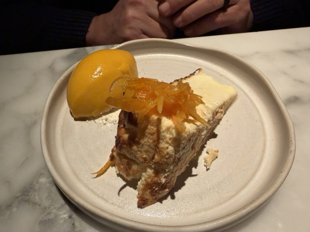 A slice of cake topped with orange zest is served on a plate alongside a scoop of bright yellow sorbet. A persons hands are visible in the background on the marble table.