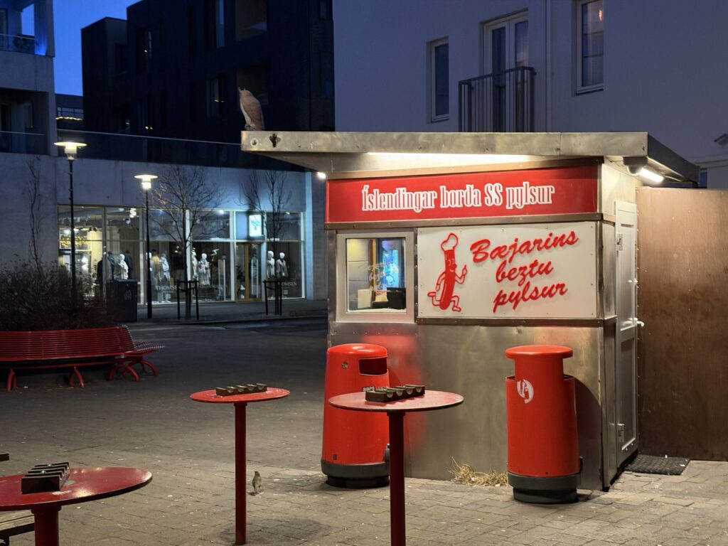 A small hot dog stand with bright red accents, Bæjarins beztu pylsur, sits on a quiet street at dusk, with empty red tables and benches nearby. Modern buildings and mannequins in store windows are in the background.