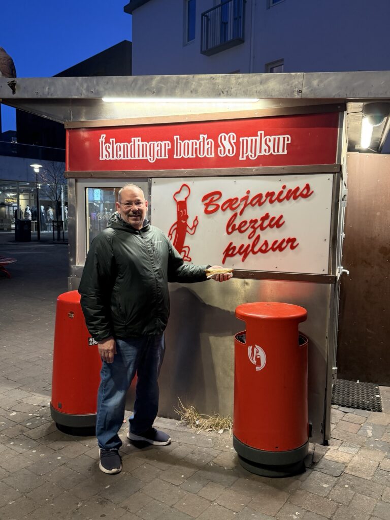 A man in a dark jacket and jeans stands smiling in front of the famous Bæjarins Beztu Pylsur hot dog stand in Reykjavik, Iceland, pointing at the sign. It is evening with streetlights on and buildings in the background.