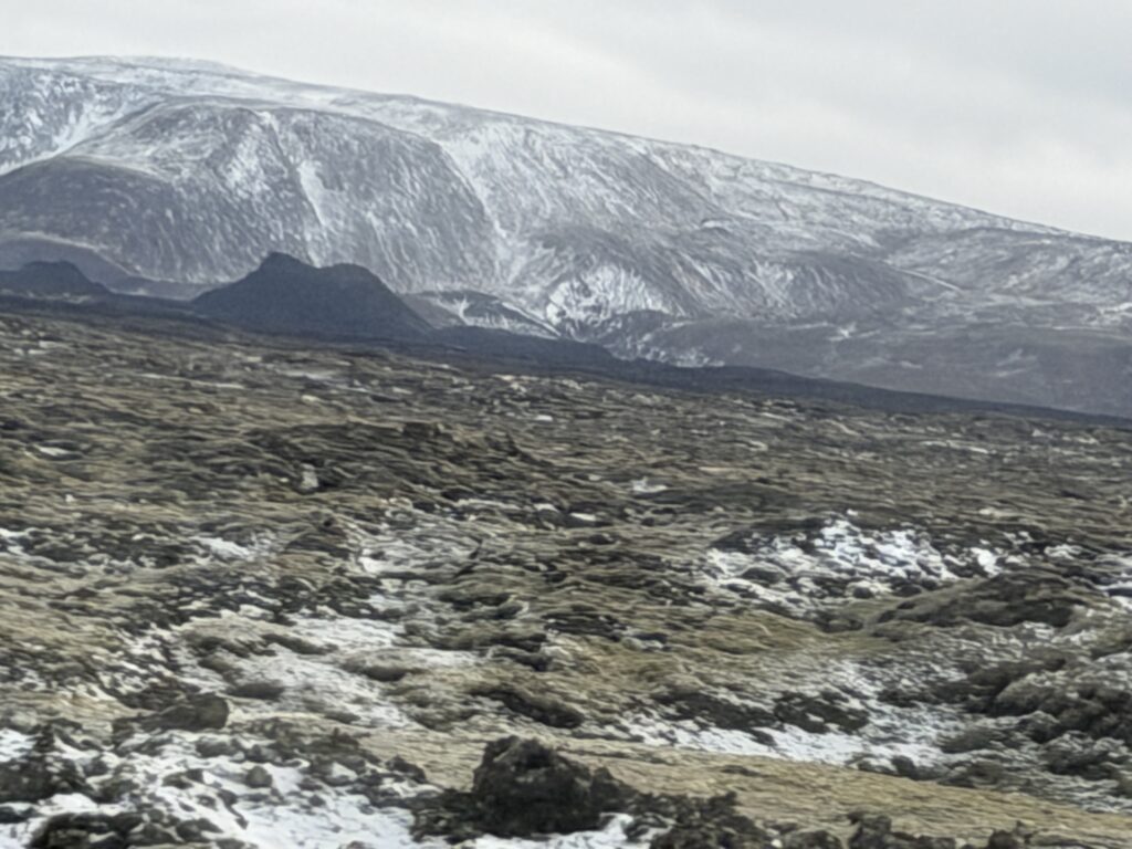 A rugged landscape with moss-covered lava rocks and patches of snow in the foreground, set against distant snow-capped mountains under a cloudy sky.