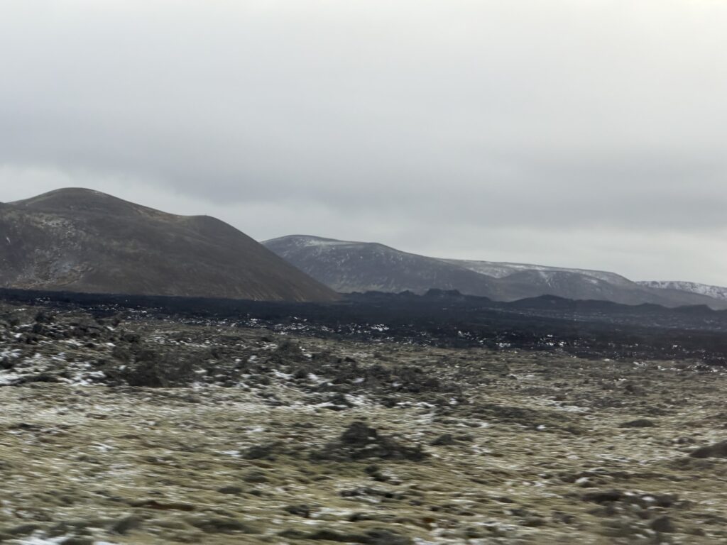 Rocky, moss-covered ground stretches toward distant, dark volcanic hills under a cloudy, gray sky, with patches of snow scattered across the landscape.