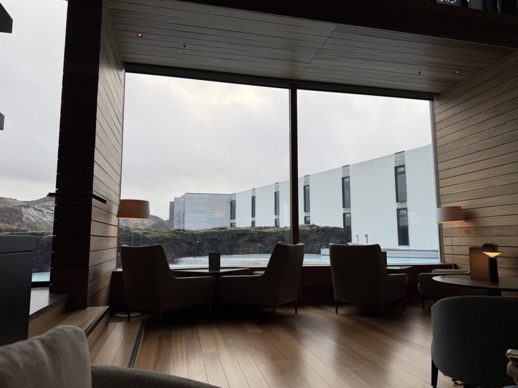 Modern lounge area with armchairs and small tables by large windows overlooking a pool and minimalist white buildings under a cloudy sky; wood floors and walls create a warm atmosphere.