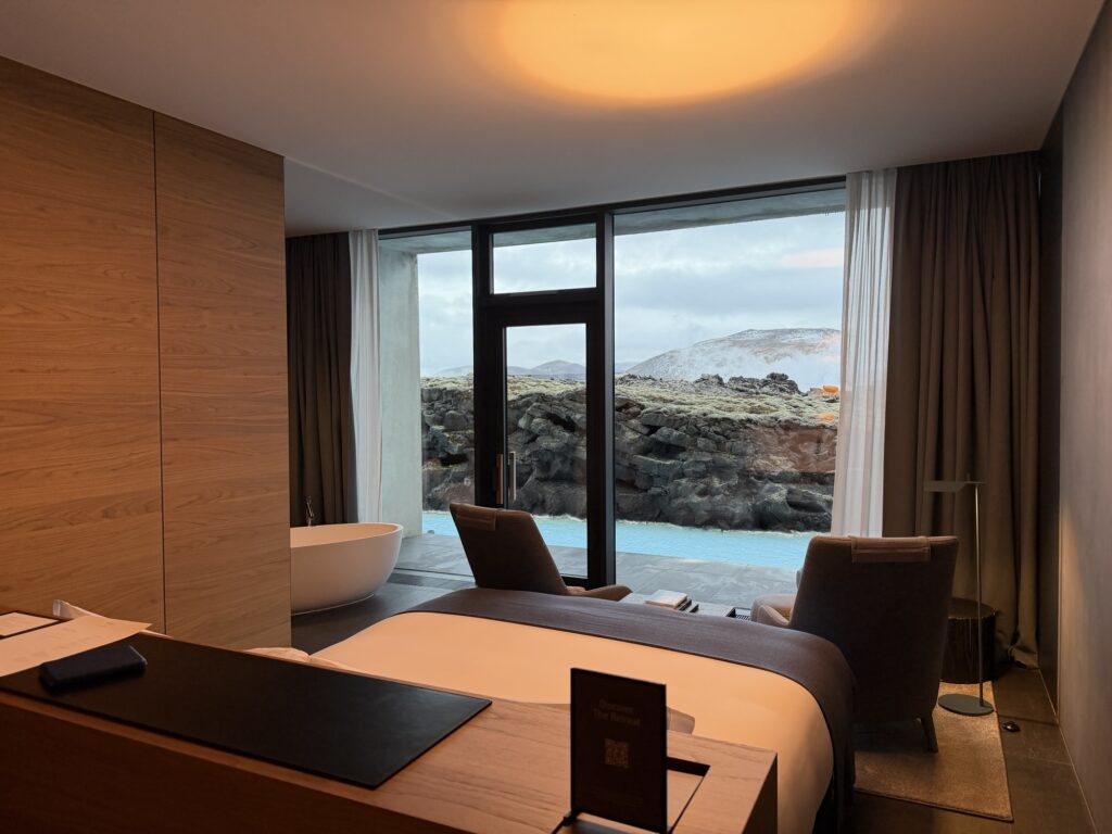 A modern hotel room with a large bed, two armchairs, a freestanding bathtub, and floor-to-ceiling windows offering a view of a rocky landscape and a pool outside.