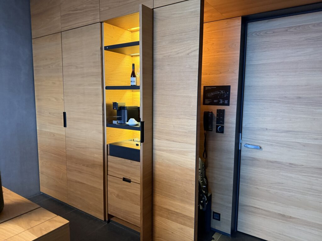 Modern wooden cabinets with built-in shelves featuring wine, glasses, a coffee maker, and cups, all illuminated by warm yellow lighting; a door with a control panel is on the right side.