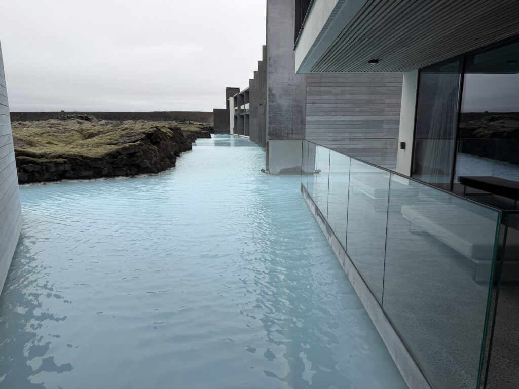 A modern building with glass railings overlooks a milky blue geothermal lagoon, surrounded by dark volcanic rocks under an overcast sky.