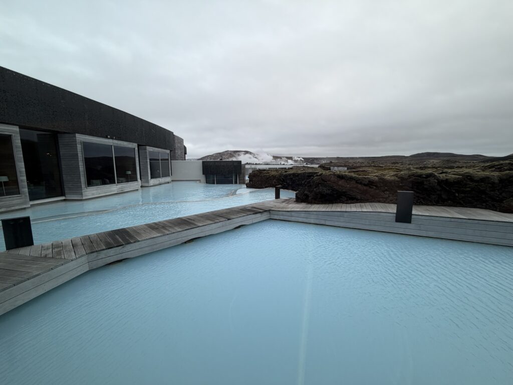 Modern buildings with large windows border a light blue geothermal pool, surrounded by volcanic rocks under a cloudy sky. Steam rises in the distance, blending with the rugged landscape.
