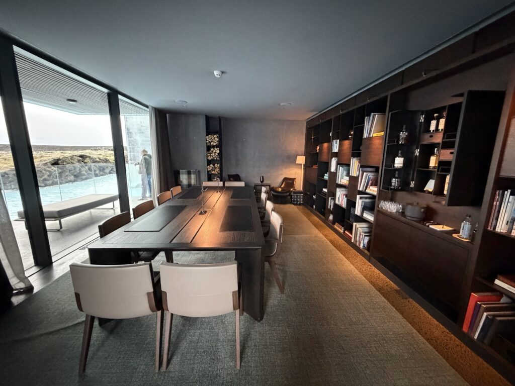 A modern dining and living area with a long wooden table, white chairs, built-in dark shelves filled with books and decor, and large windows overlooking an outdoor pool and natural landscape.