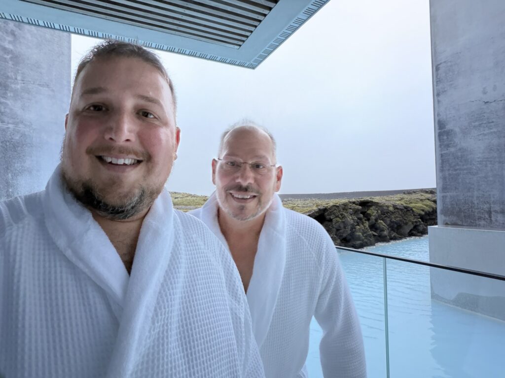 Two men in white bathrobes smile at the camera while standing on a balcony overlooking a blue geothermal pool with rocky terrain in the background.