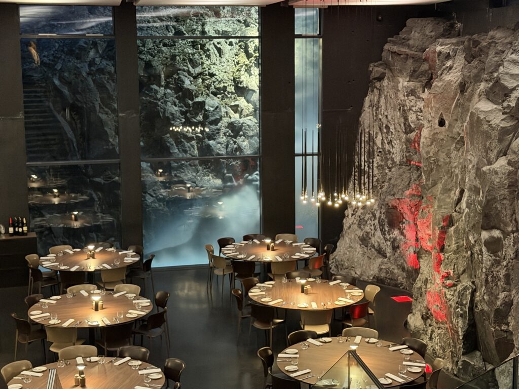 A modern restaurant with round tables set for dining, featuring a dramatic rocky wall and a large window revealing a cave-like, illuminated rock formation outside. Warm lighting and minimalist decor create an elegant atmosphere.