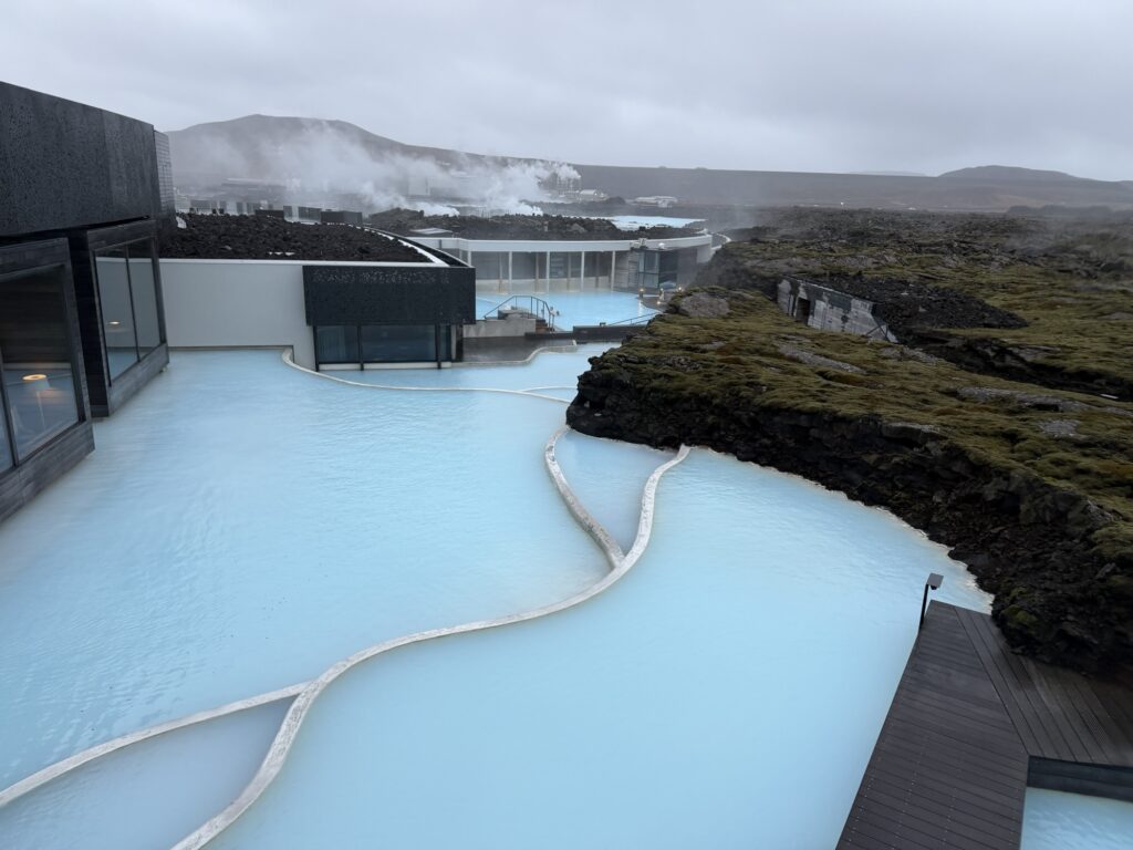 A geothermal spa with milky blue water surrounded by dark volcanic rocks and modern buildings, with steam rising in the background under a cloudy sky.