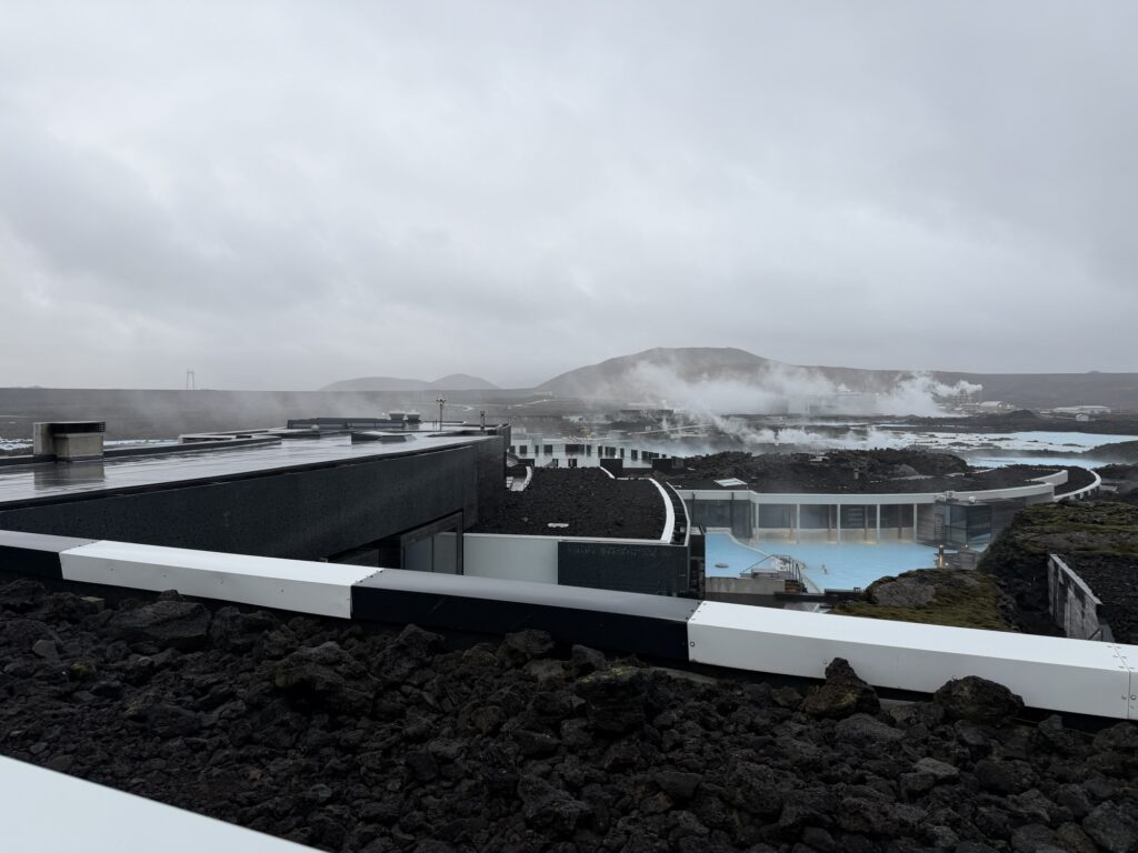A geothermal spa with steaming pools set among dark volcanic rocks under a cloudy sky, with modern buildings and distant hills visible in the background.