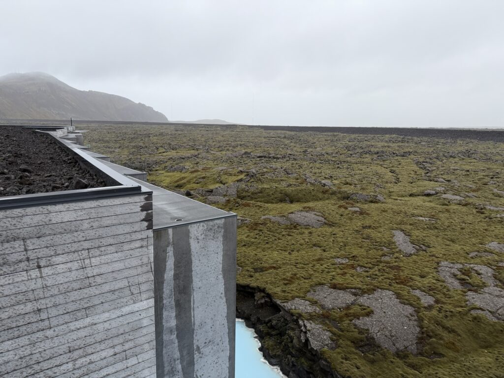 A concrete building edge overlooks a vast, moss-covered rocky landscape under a cloudy, overcast sky. A faint blue stream is visible below, and distant hills fade into the mist.