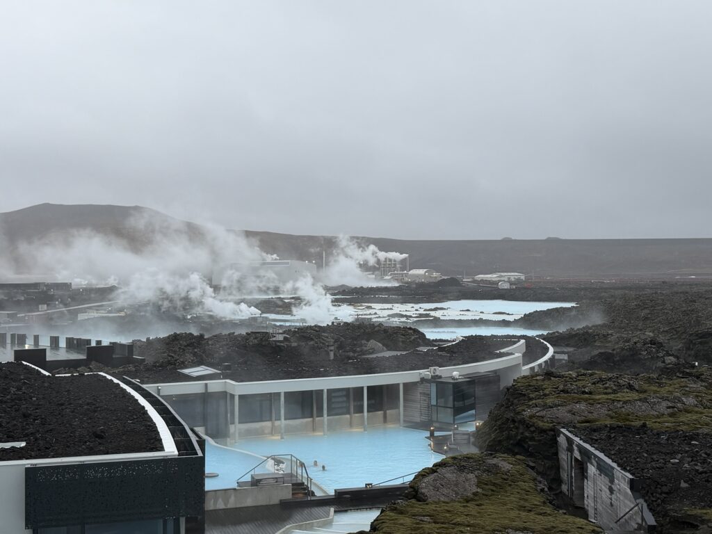 Geothermal hot springs with steaming pools set among dark volcanic rocks under a cloudy sky, with a few people bathing in the water and distant hills in the background.