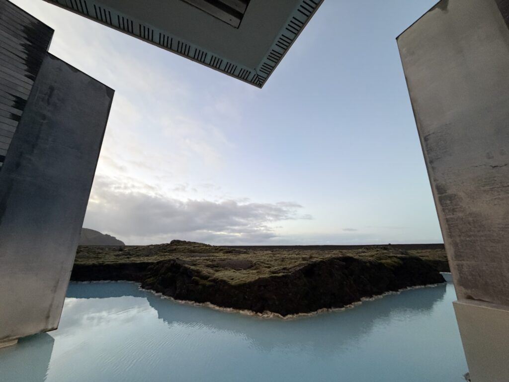Steaming blue geothermal water bordered by mossy rocks, framed by modern concrete buildings under a partly cloudy sky at sunset or sunrise.