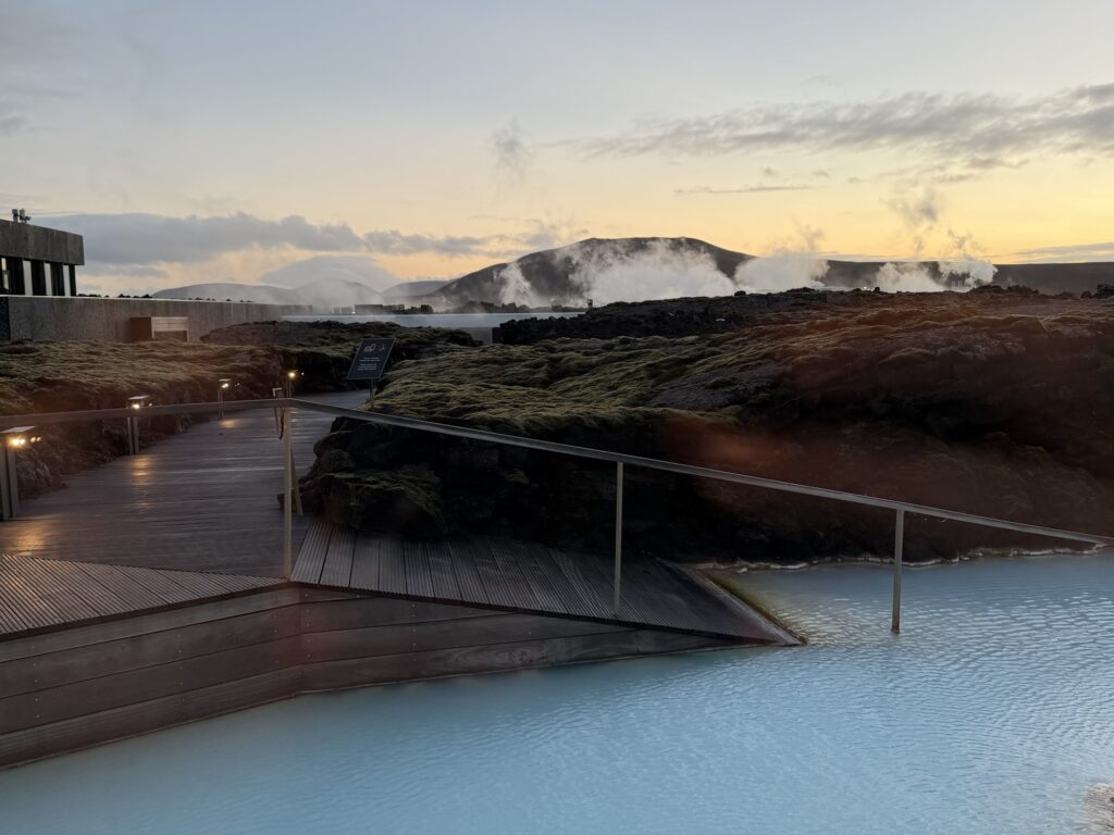 A wooden walkway with railings leads over bright blue geothermal water, surrounded by dark volcanic rocks and moss, with steam rising in the distance and a sunset sky in the background.