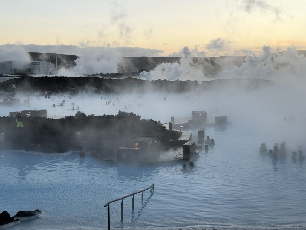 People relax in a large, steamy geothermal lagoon surrounded by dark rocks at sunset. Mist rises from the blue water, and scattered lights illuminate the scene, creating a tranquil, atmospheric setting.