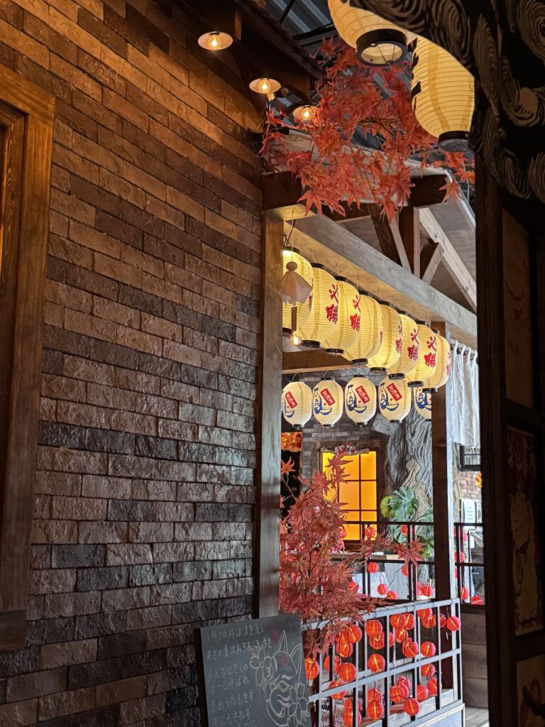 Warmly lit Japanese-style interior with brick walls, red maple leaves, hanging lanterns with Japanese characters, and a chalkboard at the entrance, creating a cozy and festive ambiance.