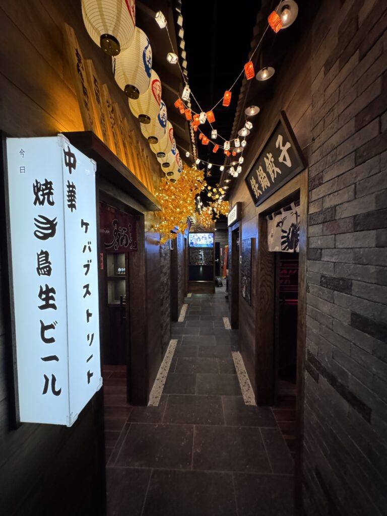 A dimly lit hallway in a Japanese restaurant, decorated with colorful hanging lanterns and signs with Japanese writing on wooden walls, leading to private dining rooms at the end.