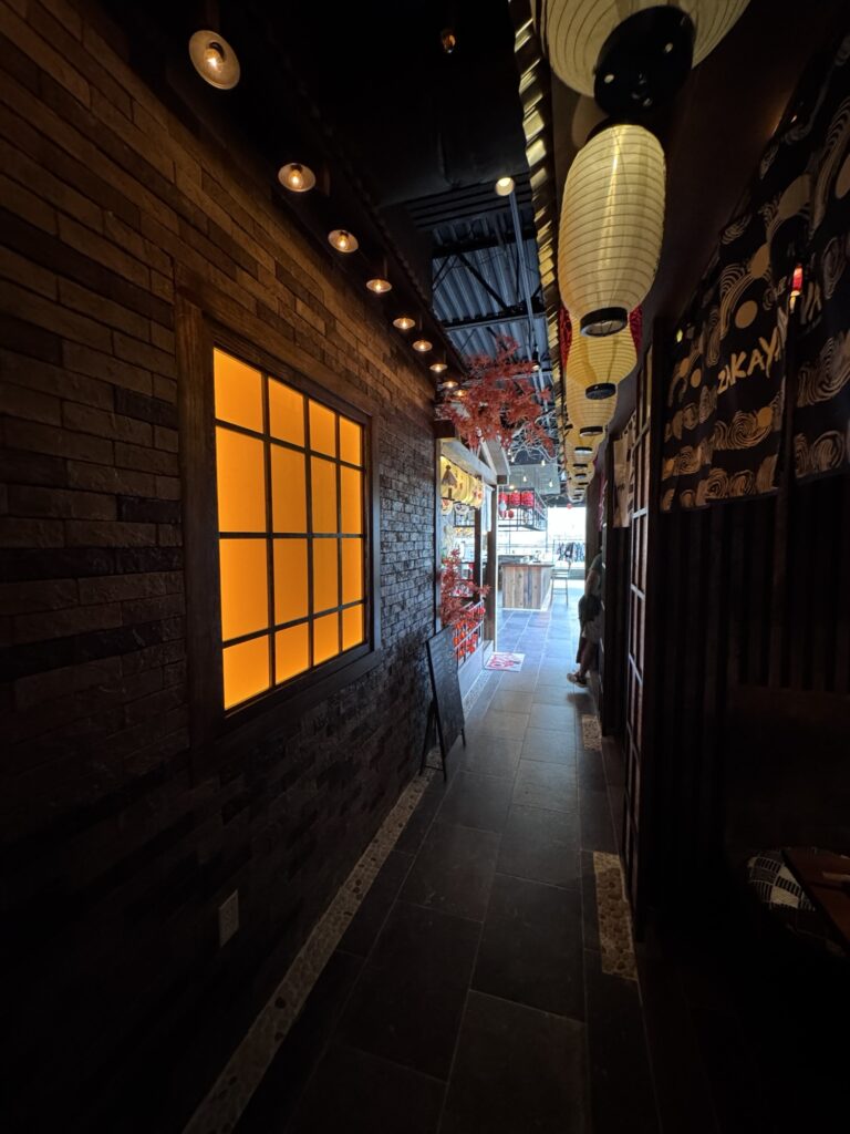 A dimly lit hallway in a Japanese-style restaurant features lanterns hanging from the ceiling, a glowing orange window on the left, and decorations leading toward a brightly lit entrance at the far end.