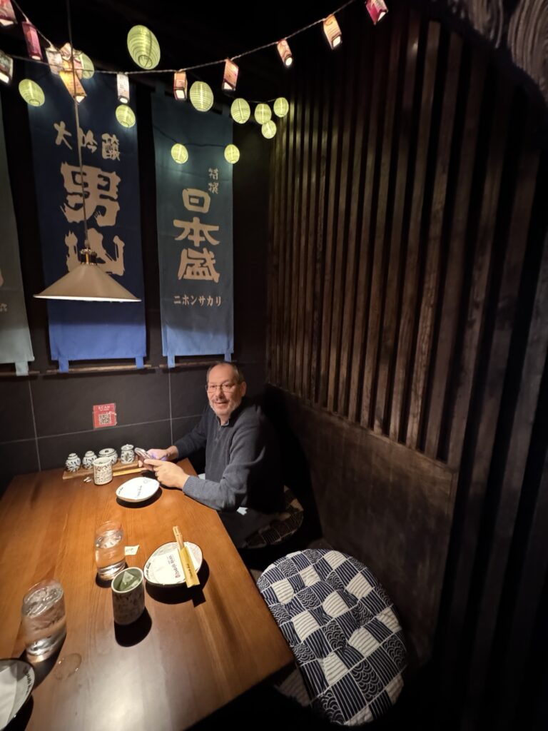 A man sits alone at a wooden table in a cozy Japanese restaurant with hanging lanterns, blue banners with Japanese writing, and patterned seat cushions. Plates, cups, and water glasses are on the table.