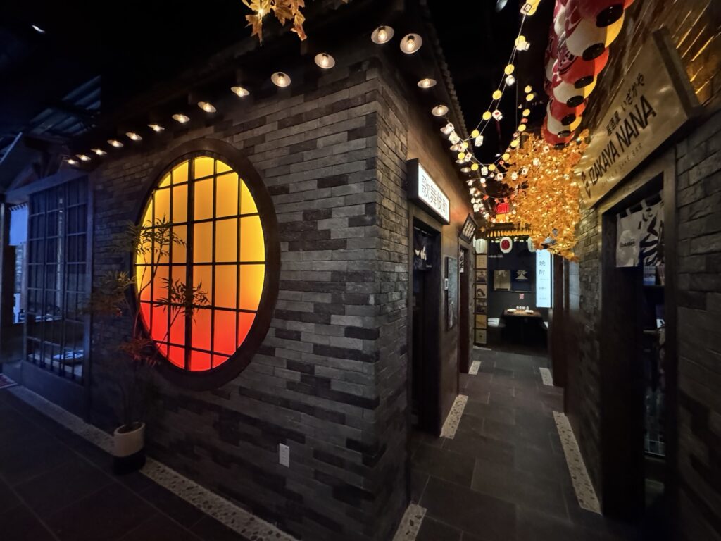 A dimly lit hallway with brick walls, round stained glass window glowing orange and yellow, hanging string lights, and a doorway under a sign reading TONKATSU NANA. Decorative plants and lanterns add ambiance.