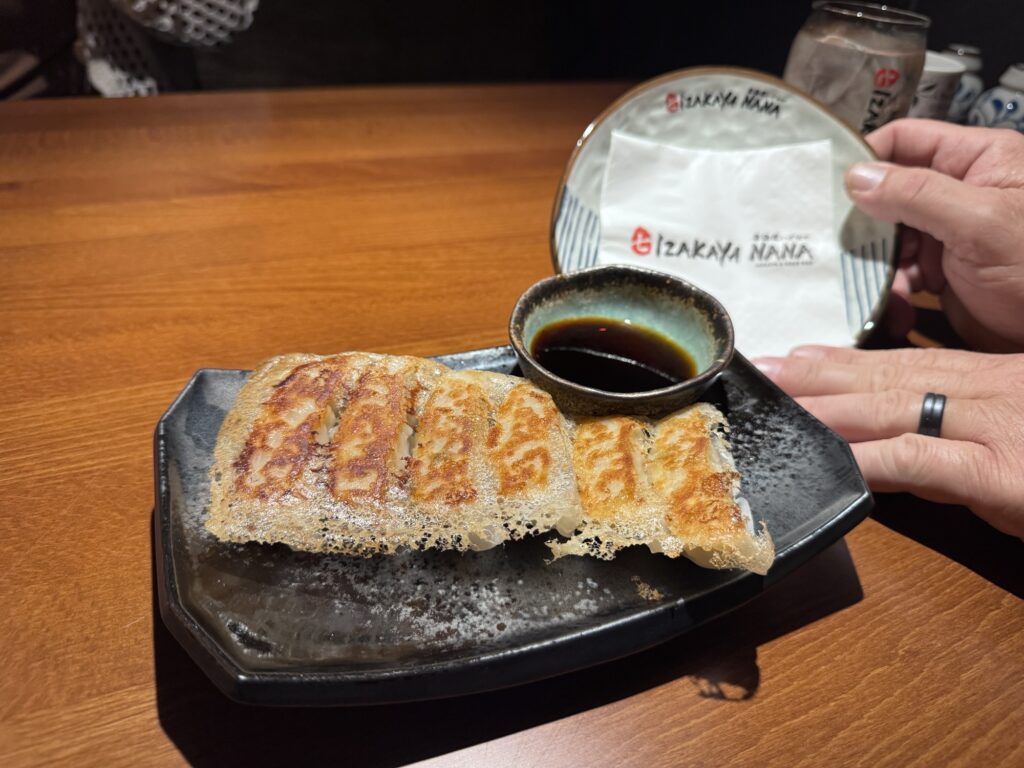 A black plate with crispy pan-fried gyoza served alongside a small dish of dipping sauce on a wooden table. A hand holds a napkin and plate with the Izakaya Nana logo in the background.
