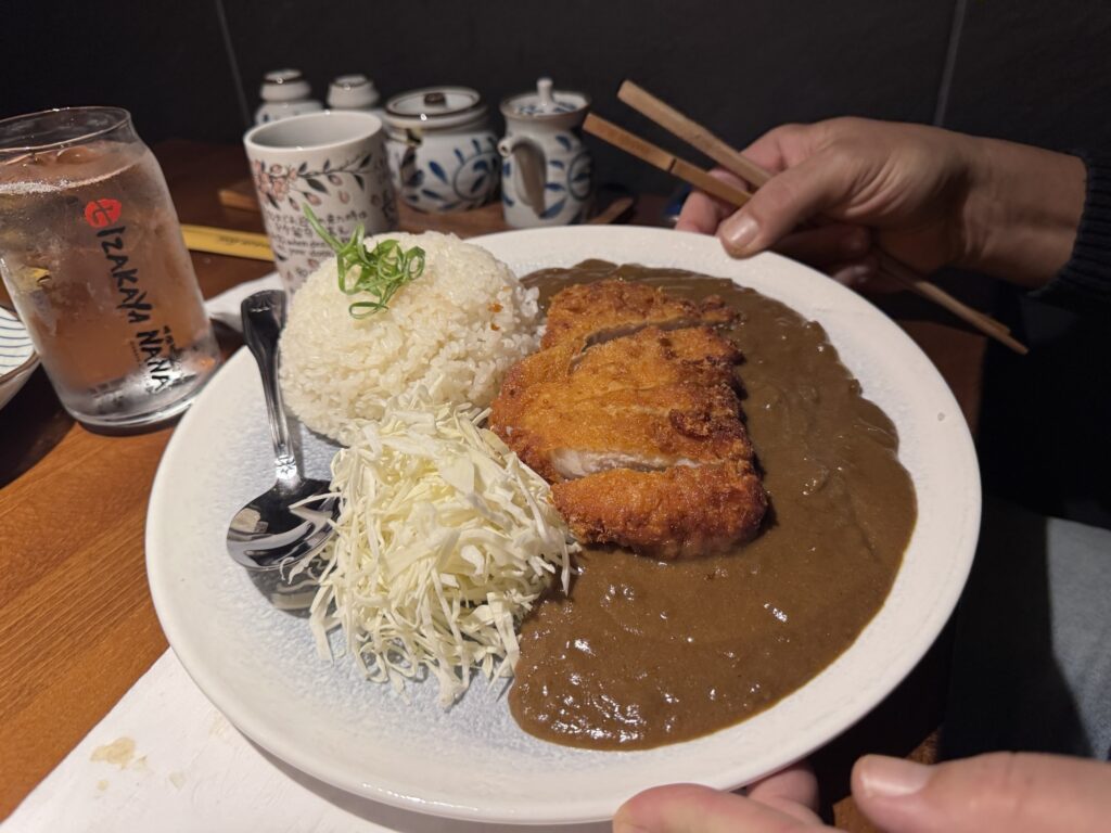 A person holds a plate with Japanese curry rice, featuring breaded fried cutlet, shredded cabbage, and a mound of steamed white rice, alongside a spoon and chopsticks. Cups and a water glass are in the background.