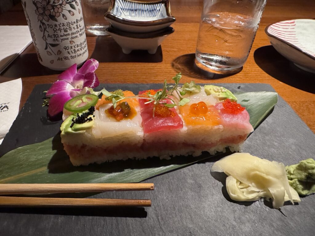 A rectangular piece of sushi topped with colorful fish, roe, avocado, herbs, and garnished with an orchid, served on a leaf with ginger and wasabi. Chopsticks and a glass of water are nearby on a wooden table.