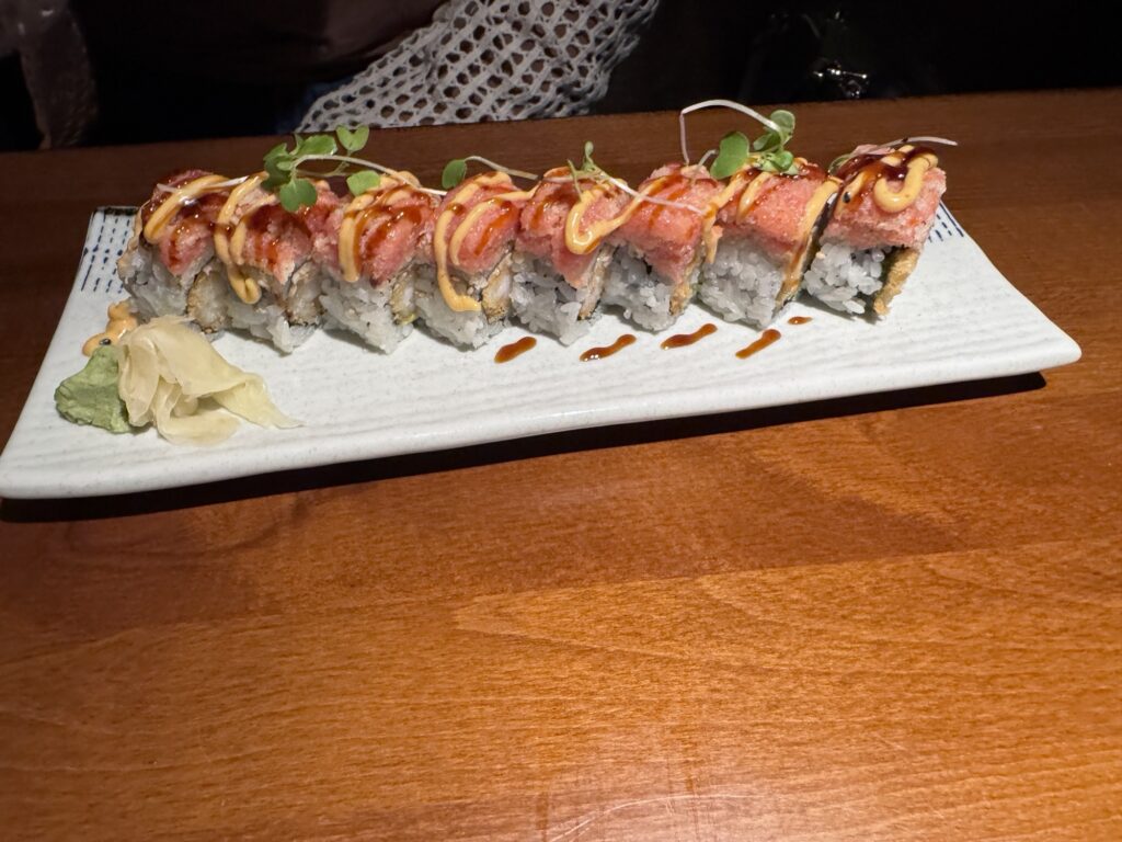 A rectangular plate of sushi rolls topped with spicy mayo, microgreens, and sauce, garnished with pickled ginger and wasabi, on a wooden table.