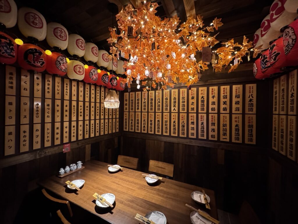 A cozy Japanese restaurant room with wooden walls decorated with menu boards, a wooden table set for four, red and white paper lanterns, and a glowing tree-like light fixture with orange leaves hanging from the ceiling.