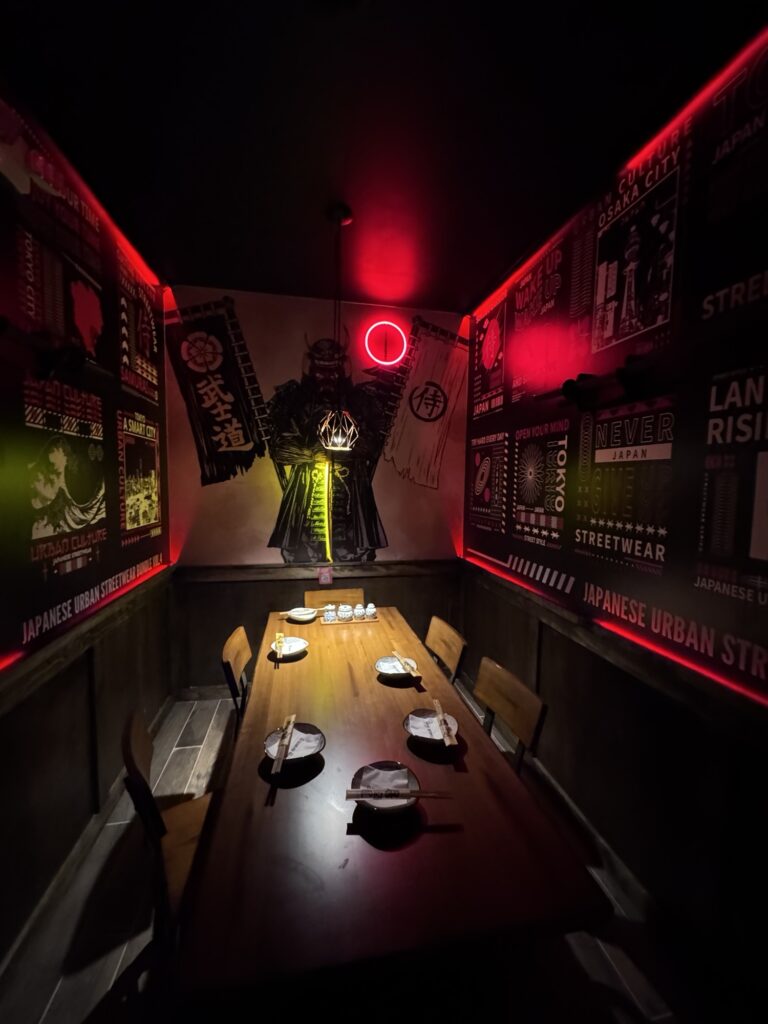 A dimly lit restaurant room features a wooden table set for six. Red neon lighting highlights Japanese urban street art and a samurai figure on the back wall, creating a dramatic atmosphere.