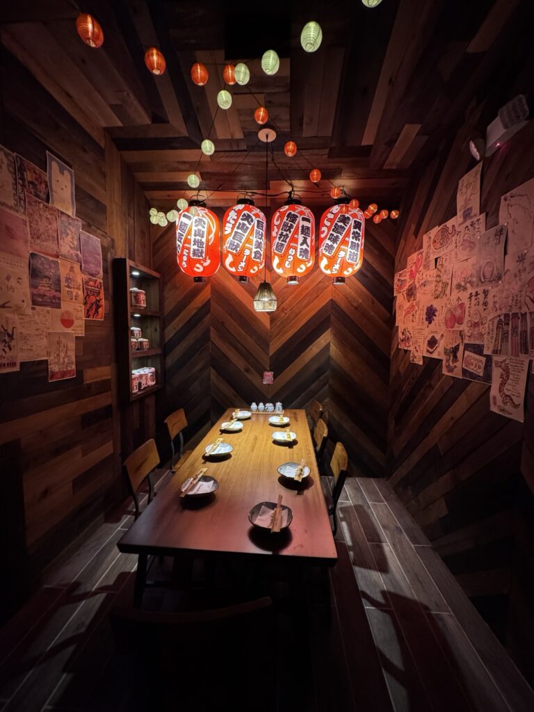 A cozy, dimly lit dining room with a wooden table set for six, Japanese lanterns hanging above, and colorful posters covering the wood-paneled walls. Warm, inviting atmosphere with a blend of modern and traditional decor.