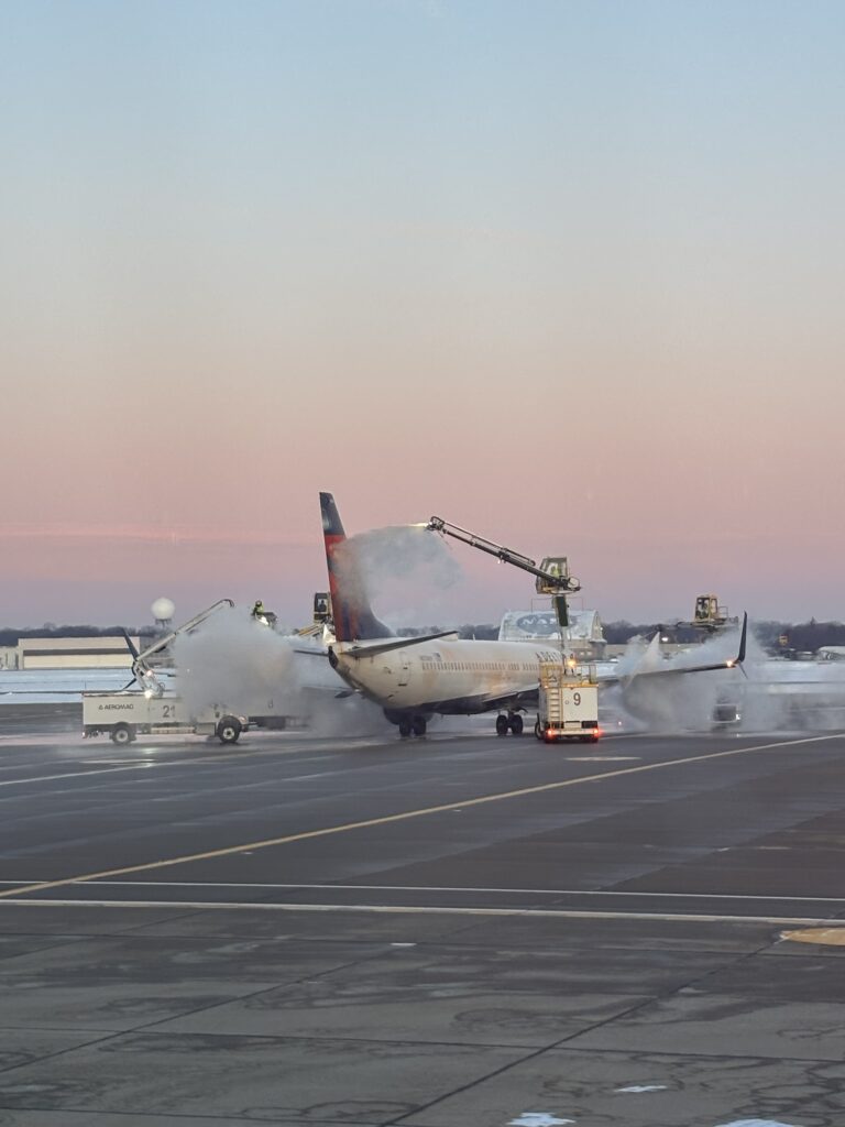 An airplane on an airport tarmac is being sprayed with de-icing fluid by multiple service vehicles under a pastel-colored evening sky. Steam or mist surrounds the aircraft as the process takes place.
