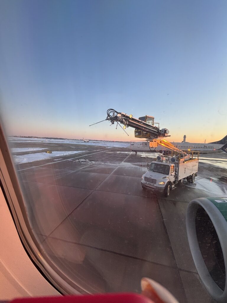 A view from an airplane window shows a de-icing truck spraying fluid onto the aircraft’s wing at sunrise, with a clear sky, snow on the ground, and airport runway in the background.