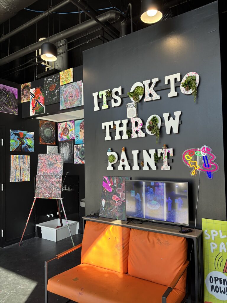 A colorful art studio with an orange couch, a black wall displaying “ITS OK TO THROW PAINT,” vibrant abstract art on the walls, an easel with a splatter painting, and paint supplies on the floor.