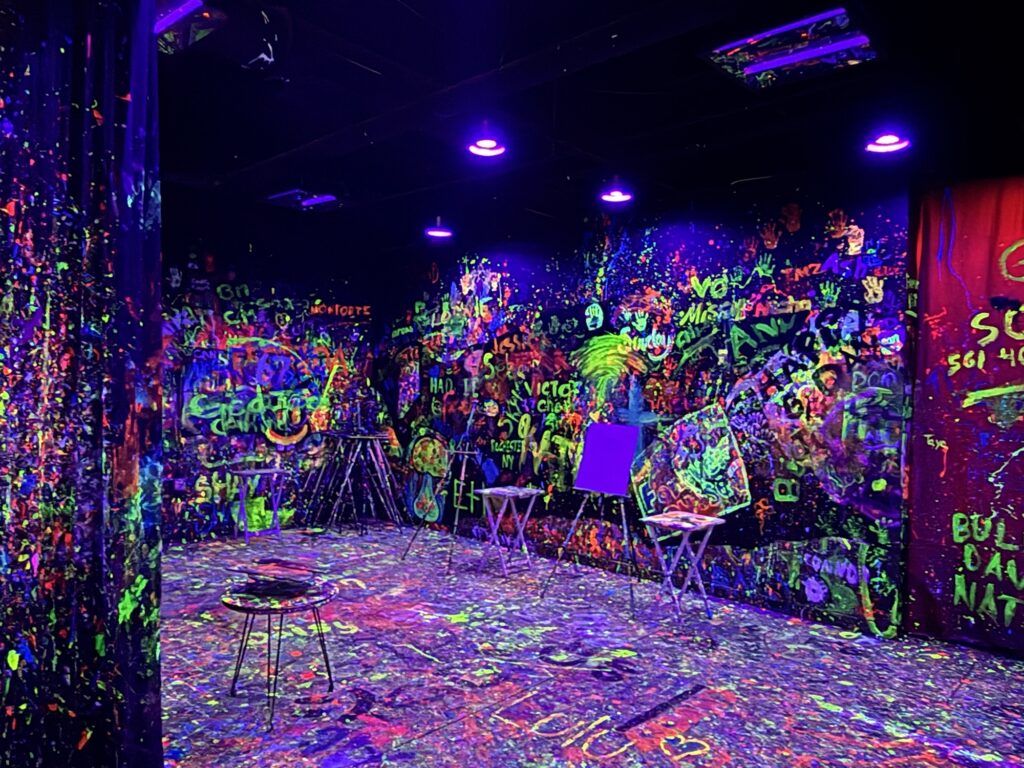 A room illuminated by black lights, with walls, floor, and easels covered in neon paint splatters and graffiti, creating a vibrant, chaotic, and colorful artistic environment.