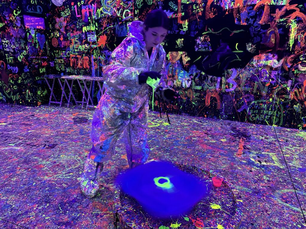 A person in a protective suit stands in a room covered in neon splatter paint, pouring glowing paint onto a spinning circular canvas under blacklight. The scene is vibrant and colorful, with art covering the walls and floor.