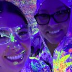 Two people smiling under blacklight, wearing clear safety goggles and protective clothing splattered with neon paint, creating a vibrant, colorful glow in the dark environment.
