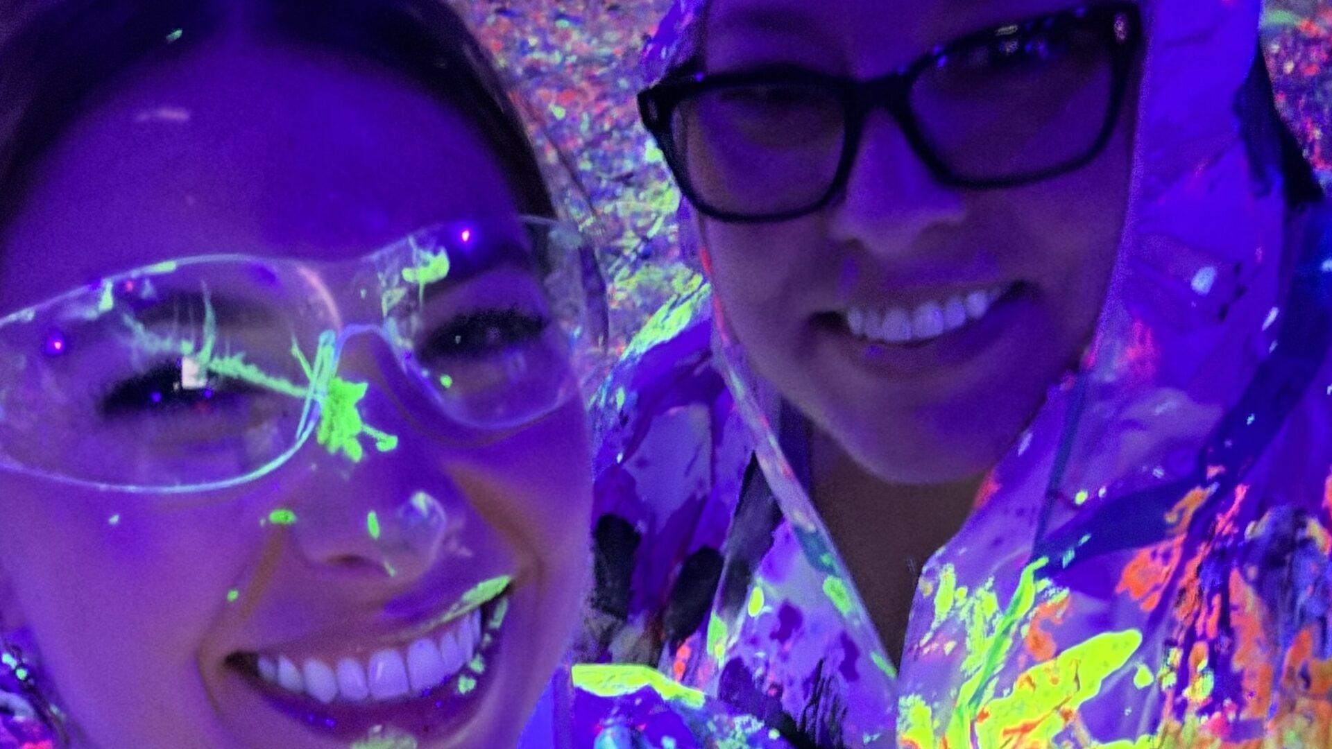 Two people smiling under blacklight, wearing clear safety goggles and protective clothing splattered with neon paint, creating a vibrant, colorful glow in the dark environment.